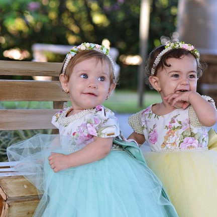 Duas bebês de vestido, olhando para câmera. Uma delas sorri enquanto morde sua mão.